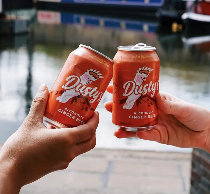 Two hands raising orange Dusty's Ginger Beer cans beside a canal