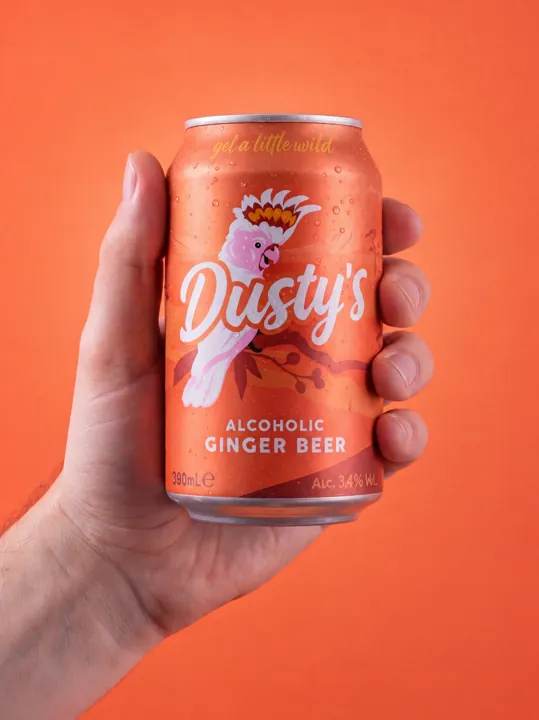 Hand holding a chilled Dusty's Ginger Beer can with condensation against a coral background