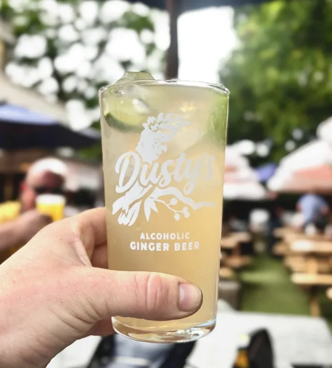 Hand holding a Dusty's Ginger Beer pint glass with lime in a beer garden