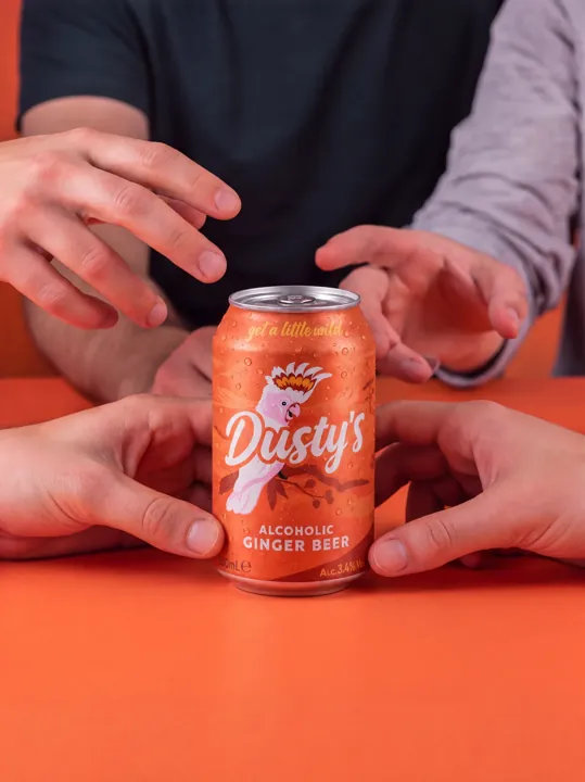Hands gathering around a chilled Dusty's Ginger Beer can on a coral table